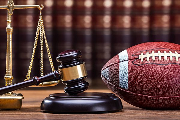 A football resting on a wooden tabletop with a judge's gavel and scales of justice.